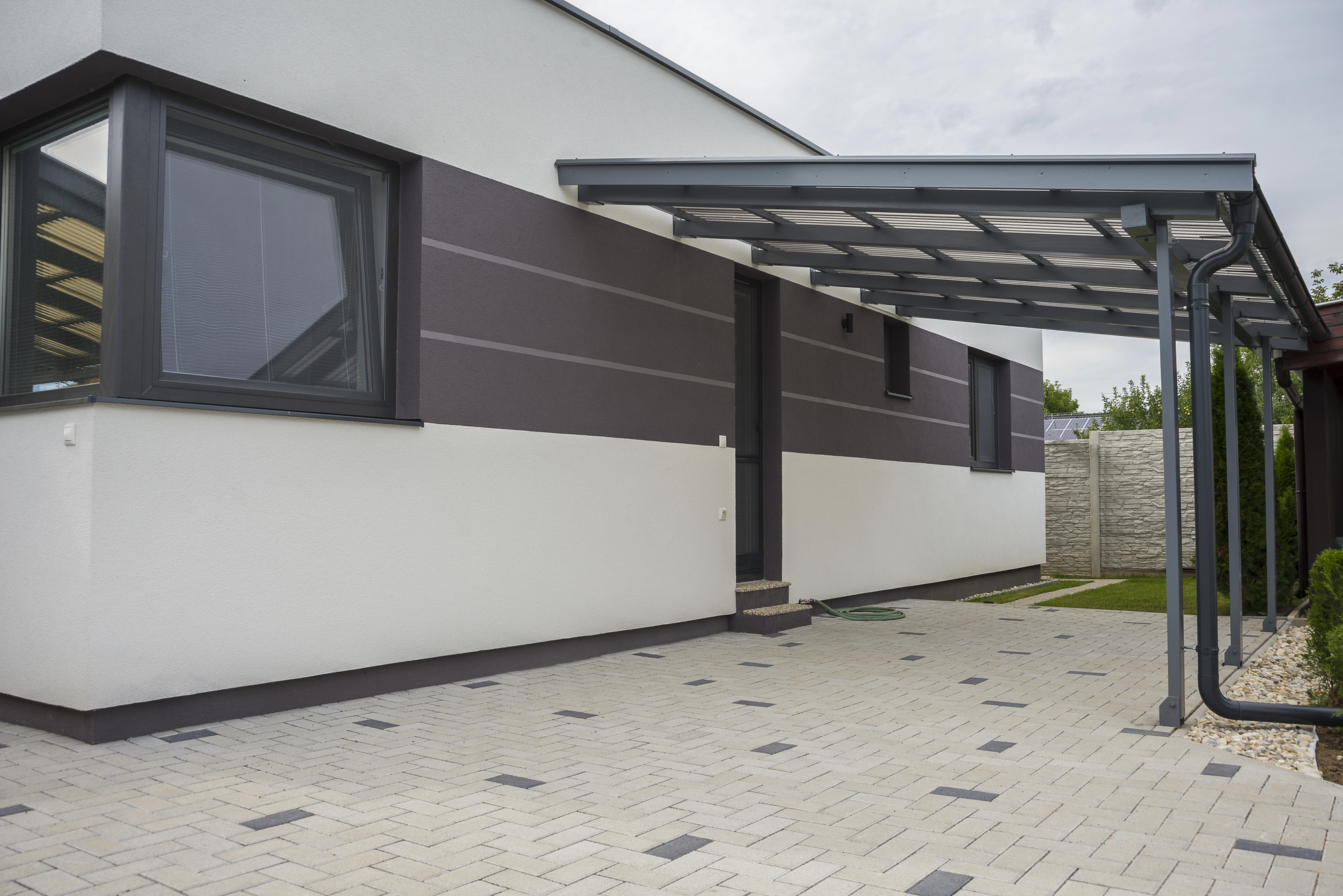 Modern house exterior with a flat roof, large windows, a covered carport, paved driveway, and a small patch of green lawn bordered by a stone fence.