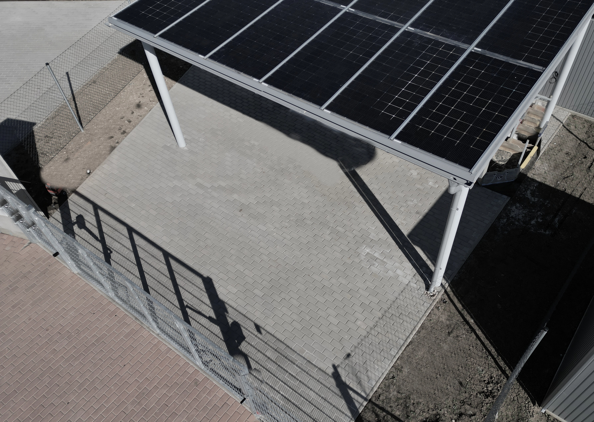 An open, paved area is partially covered by a metal structure with solar panels, bordered by a wire fence and a small patch of dirt.
