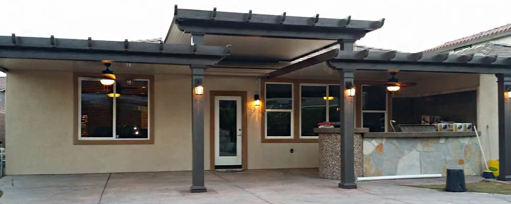 A covered backyard patio with a ceiling fan, lights, and an outdoor kitchen area with a stone bar counter attached to a beige house.