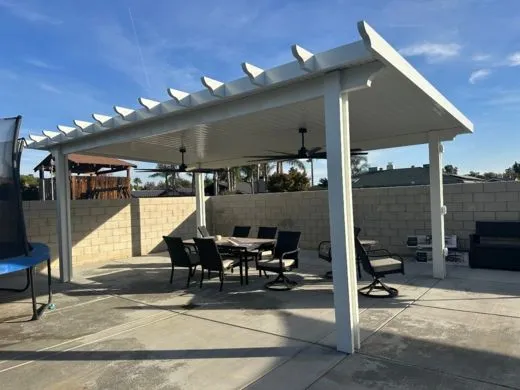 A white pergola covers an outdoor dining area with a table, chairs, and ceiling fans, set on a concrete patio near a block wall.