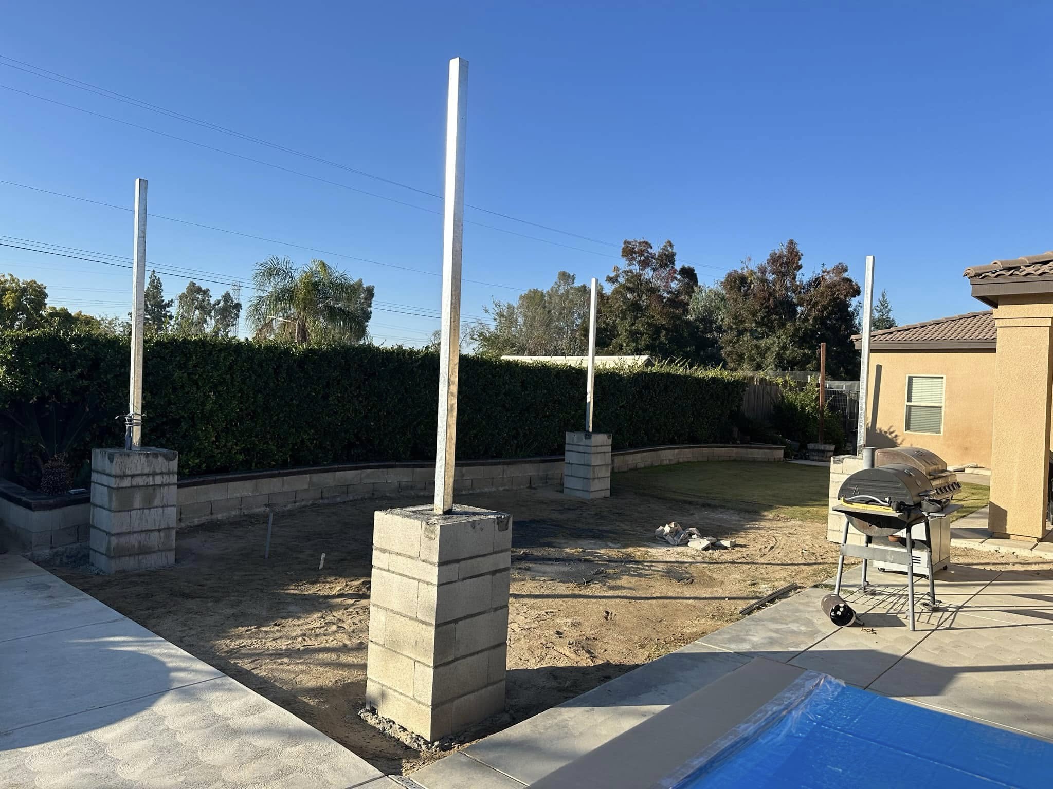 A backyard with unfinished construction shows several concrete block bases with white vertical posts, surrounded by grass, a hedge, and patio area with a grill.