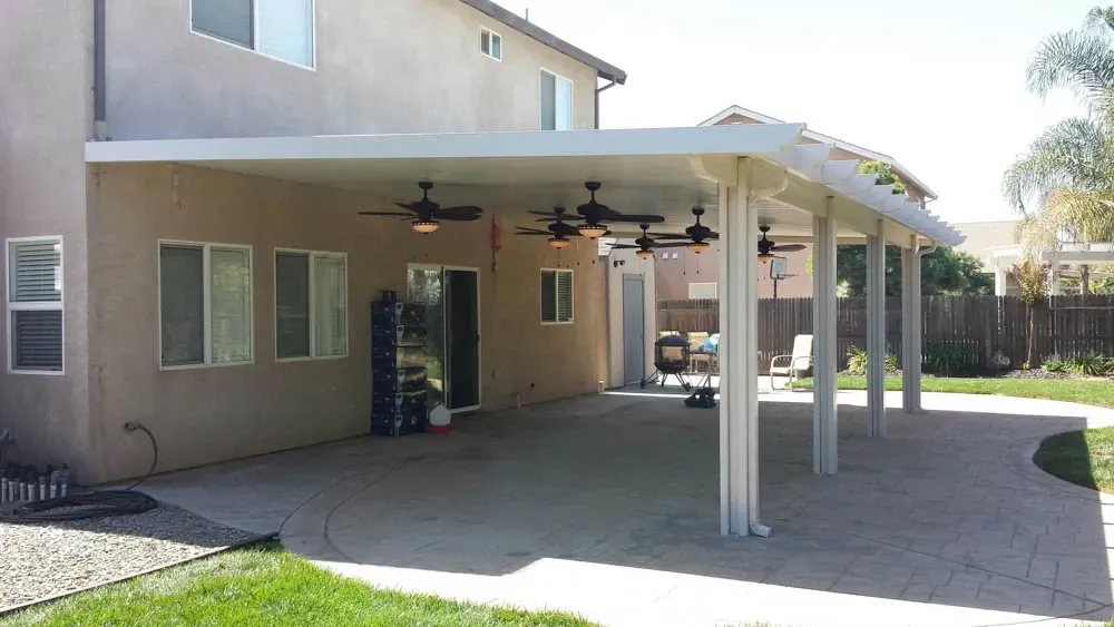 Covered backyard patio attached to a house, featuring ceiling fans, a paved floor, and some outdoor furniture in the background.