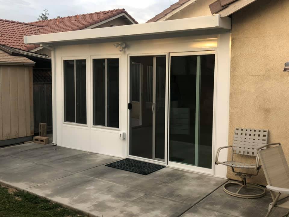 A small enclosed patio with large windows and a sliding glass door, attached to a house with a concrete patio and an outdoor chair nearby.
