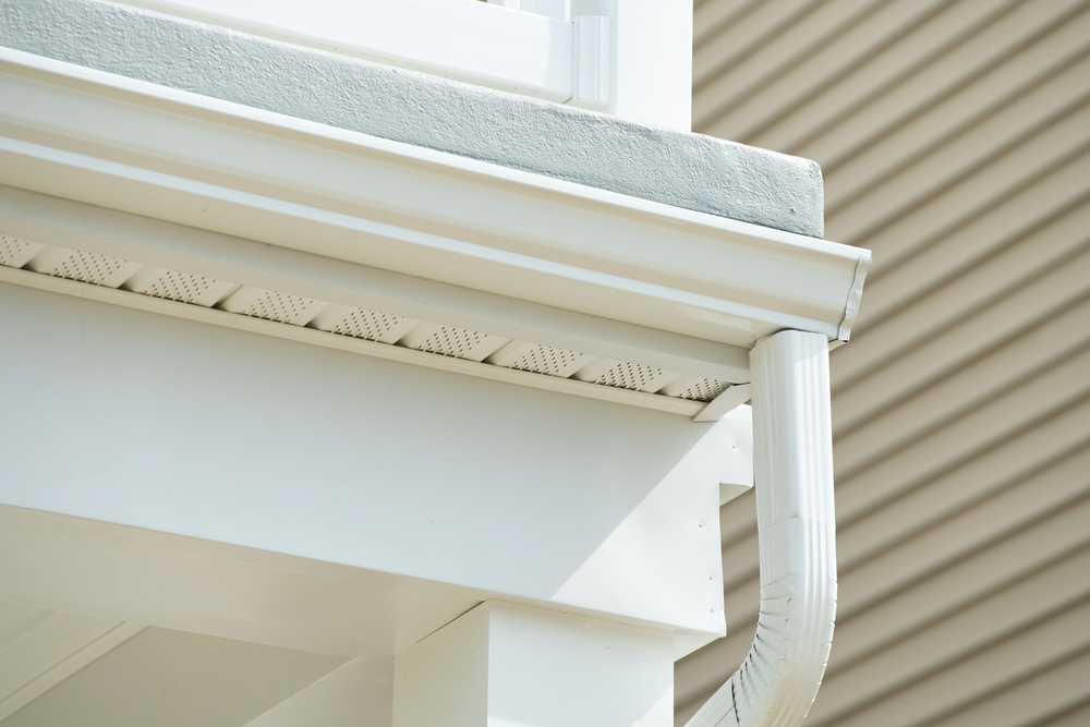 Close-up view of a white house roof edge, showing perforated soffit panels, a white gutter, and a downspout against beige siding in the background.