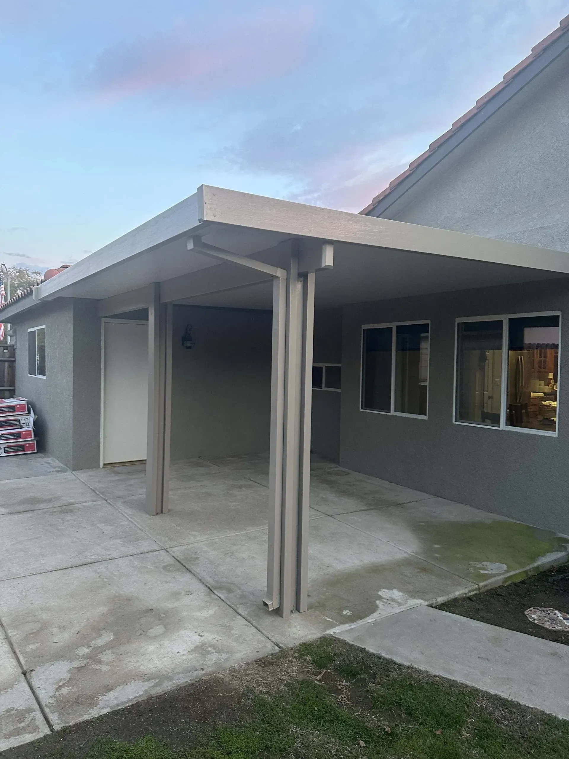 A covered patio with two supporting columns attached to a gray house, next to windows and a door, on a concrete surface.