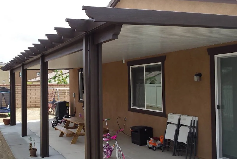 Covered backyard patio with brown posts and beams, picnic table, children’s bikes, stacked chairs, and a tan stucco house with sliding and standard windows.