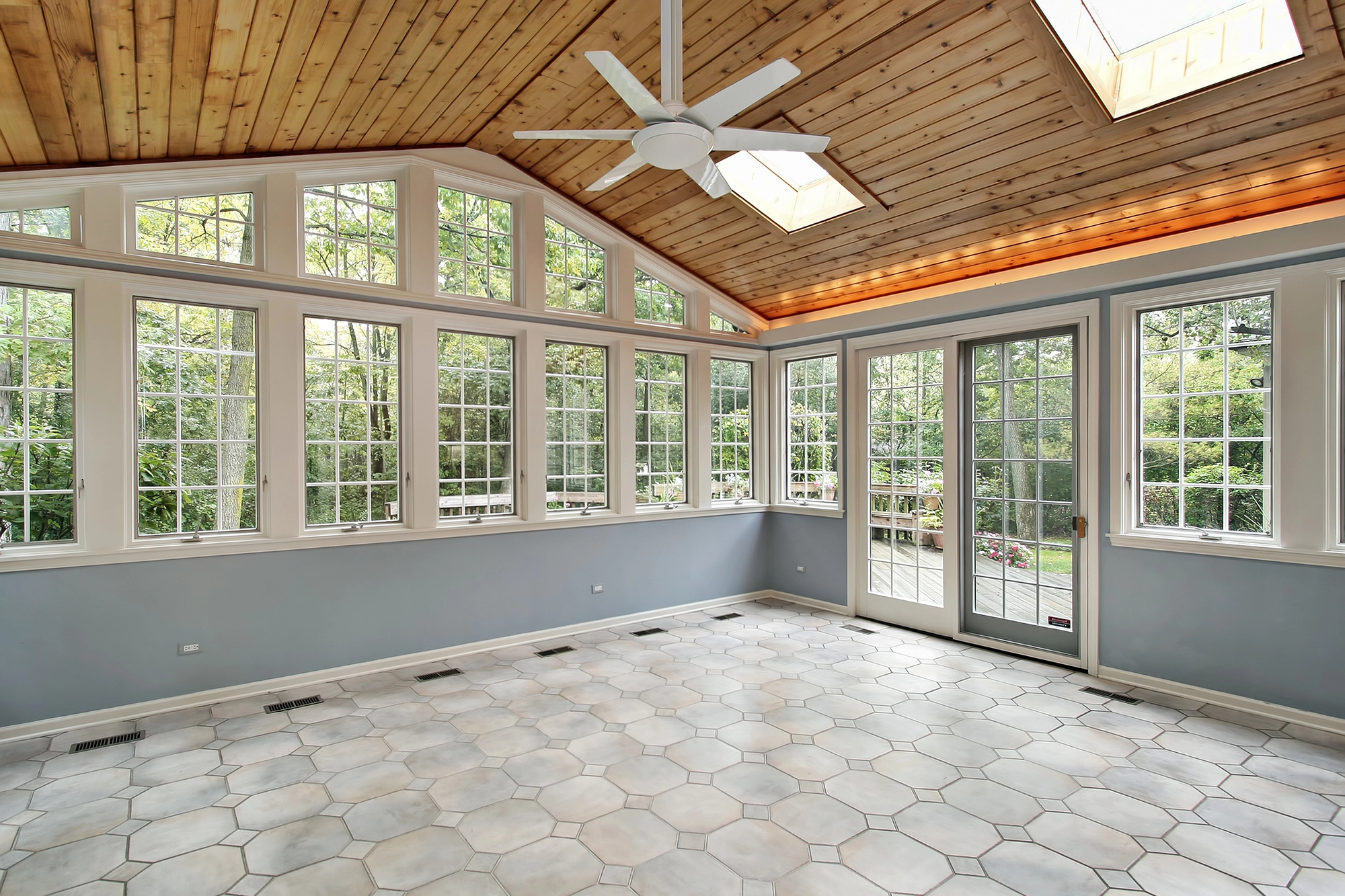 Bright sunroom with large windows, a wood-paneled ceiling, skylights, ceiling fan, light blue walls, and tiled floor overlooking a garden.