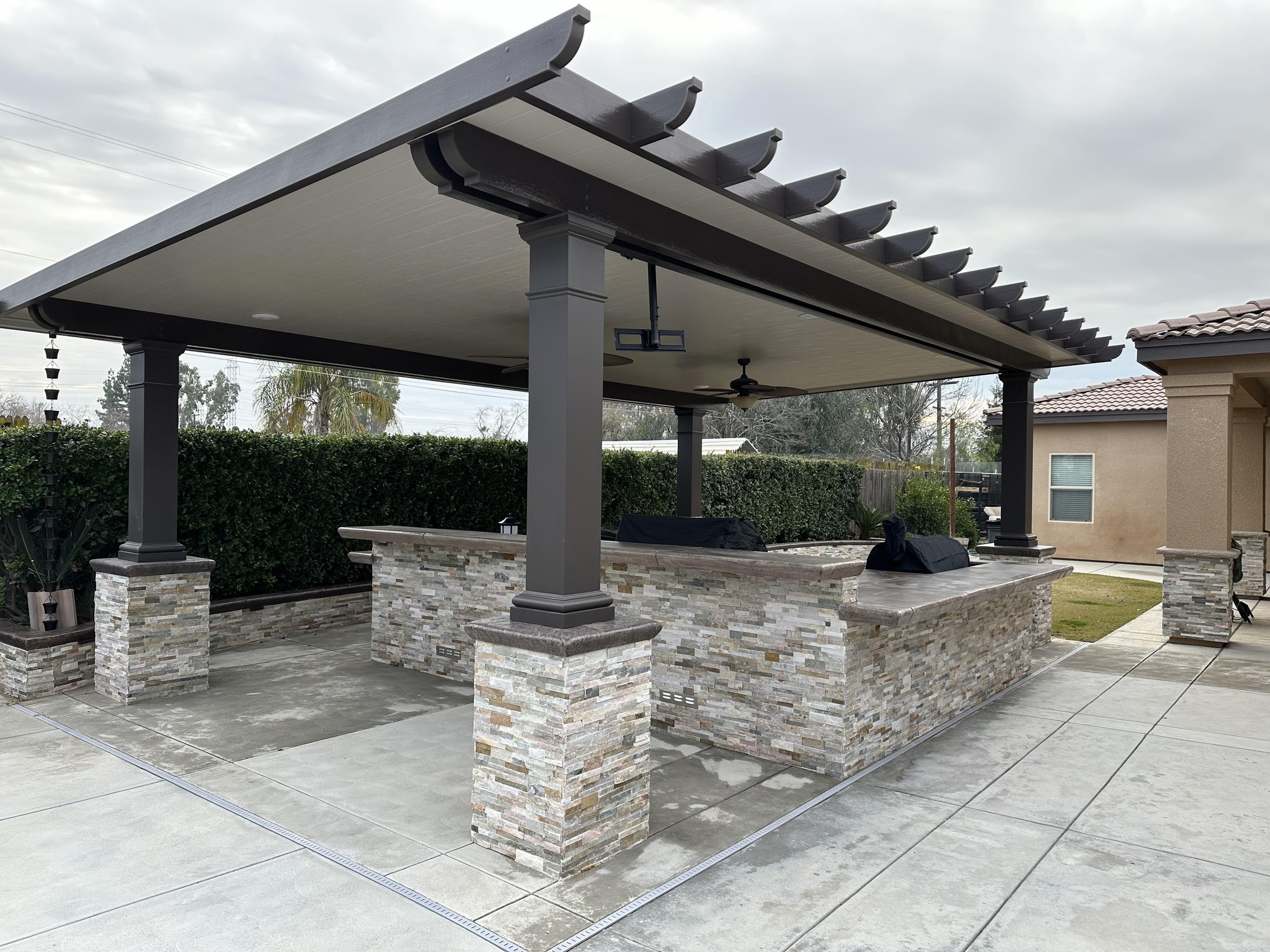 Outdoor covered patio area with a stone bar and grill setup, surrounded by hedges and adjacent to a house on a cloudy day.